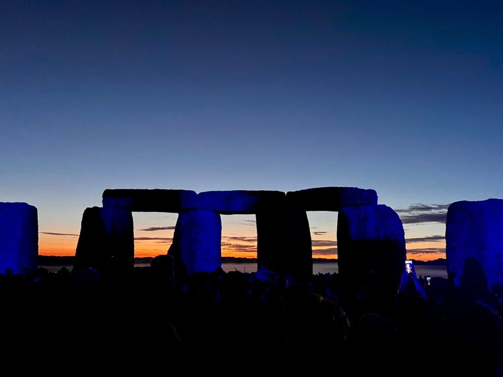 Stonehenge silhouetted against an orange and blue pre-dawn sky with the Summer Solstice crowd below.