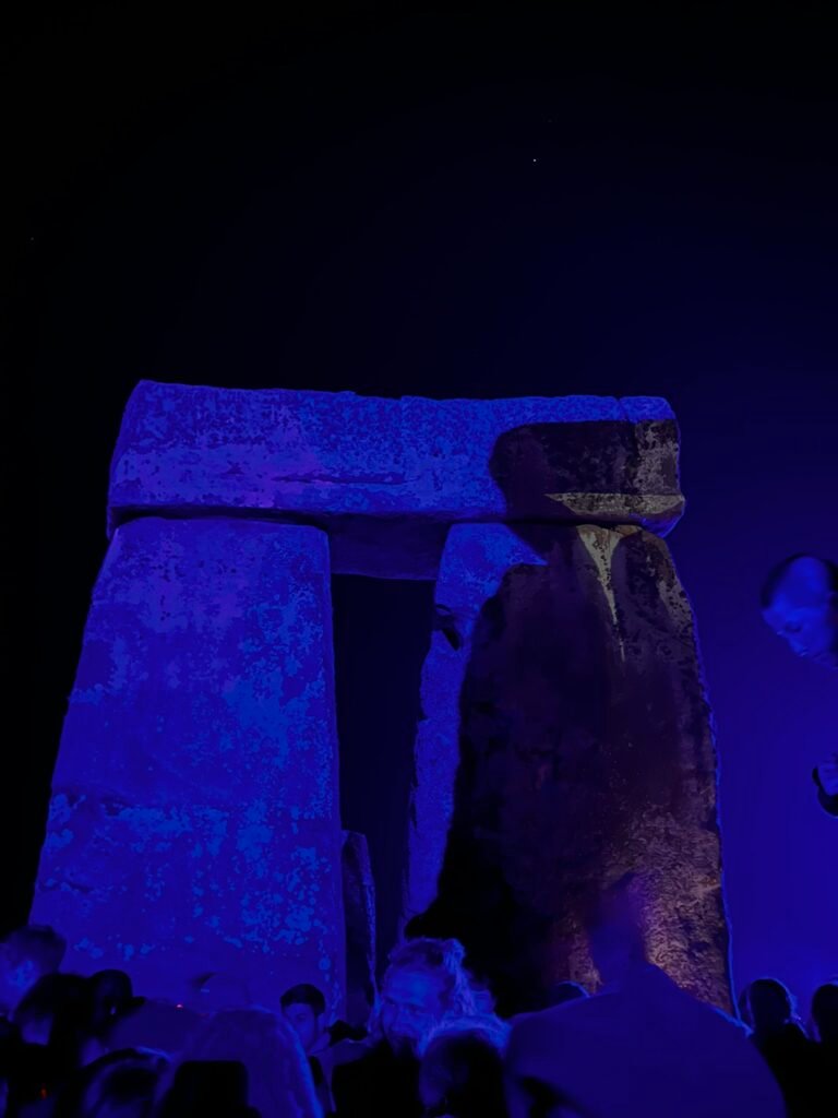 Stonehenge illuminated in blue and purple light at night with Summer Solstice visitors gathered beneath the stones.