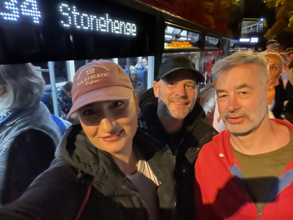 Three visitors beside a bus displaying route 334 to Stonehenge during the Summer Solstice night service.