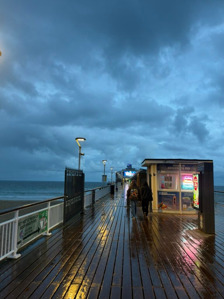 Wet wooden walkway on Bournemouth Pier reflecting colourful lights beneath a cloudy evening sky