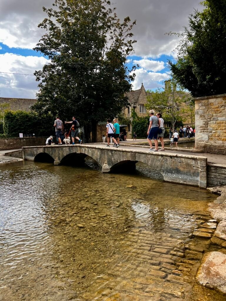 Visitors cross a low stone bridge above the shallow River Windrush.