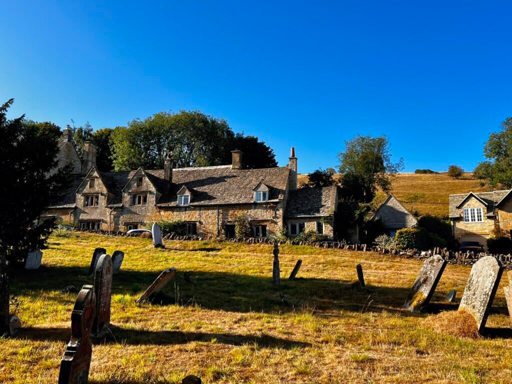 Weathered gravestones in Snowshill churchyard, with old stone cottages and golden hills beyond.