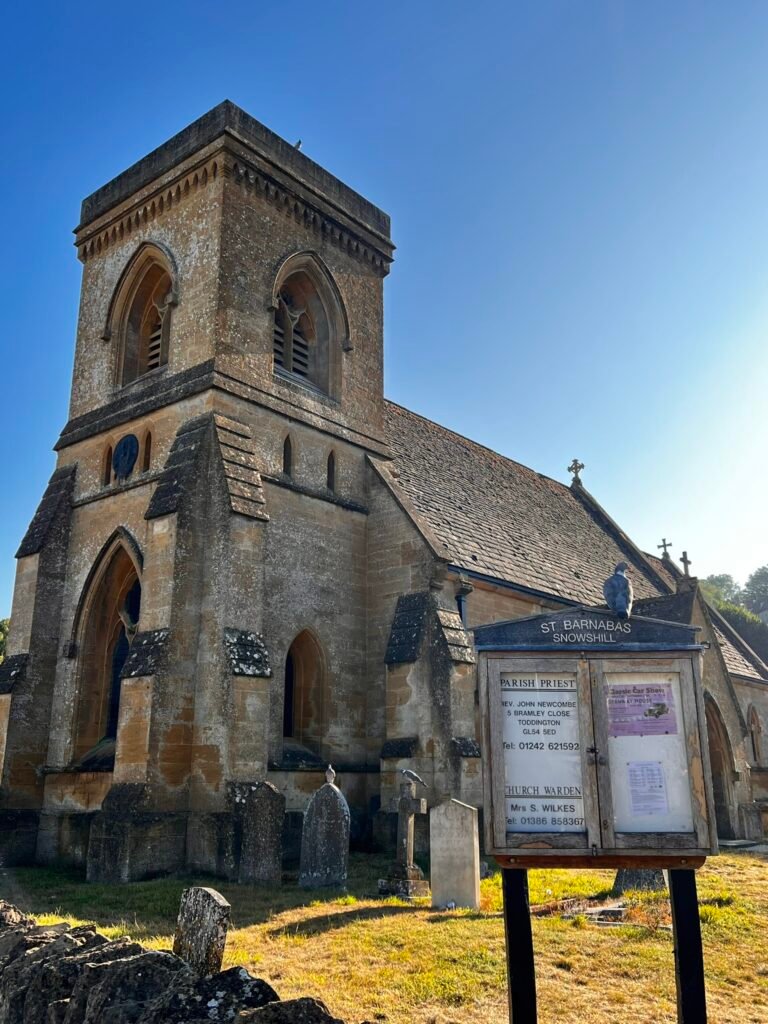 Stone tower of St Barnabas Church in Snowshill, with a village noticeboard and perched pigeon.