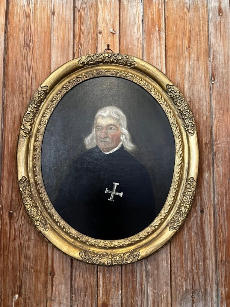 Oval portrait of a white-haired man wearing a black robe with a cross emblem at St Cross.