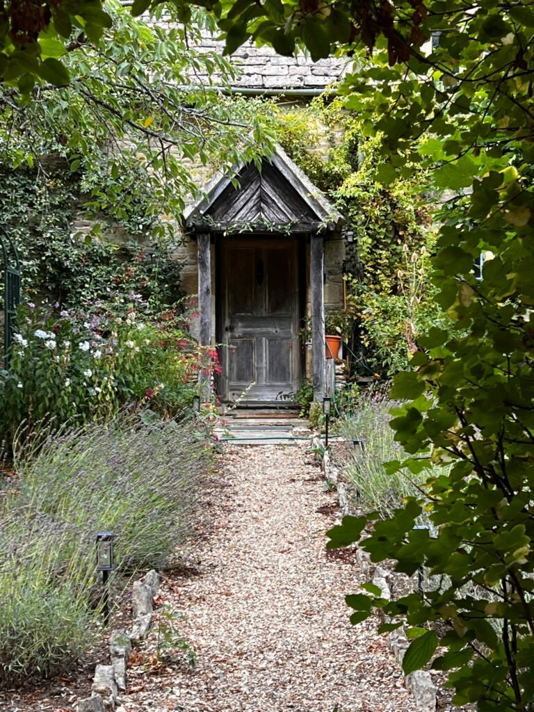 Weathered wooden cottage door reached by a garden path through leafy branches.